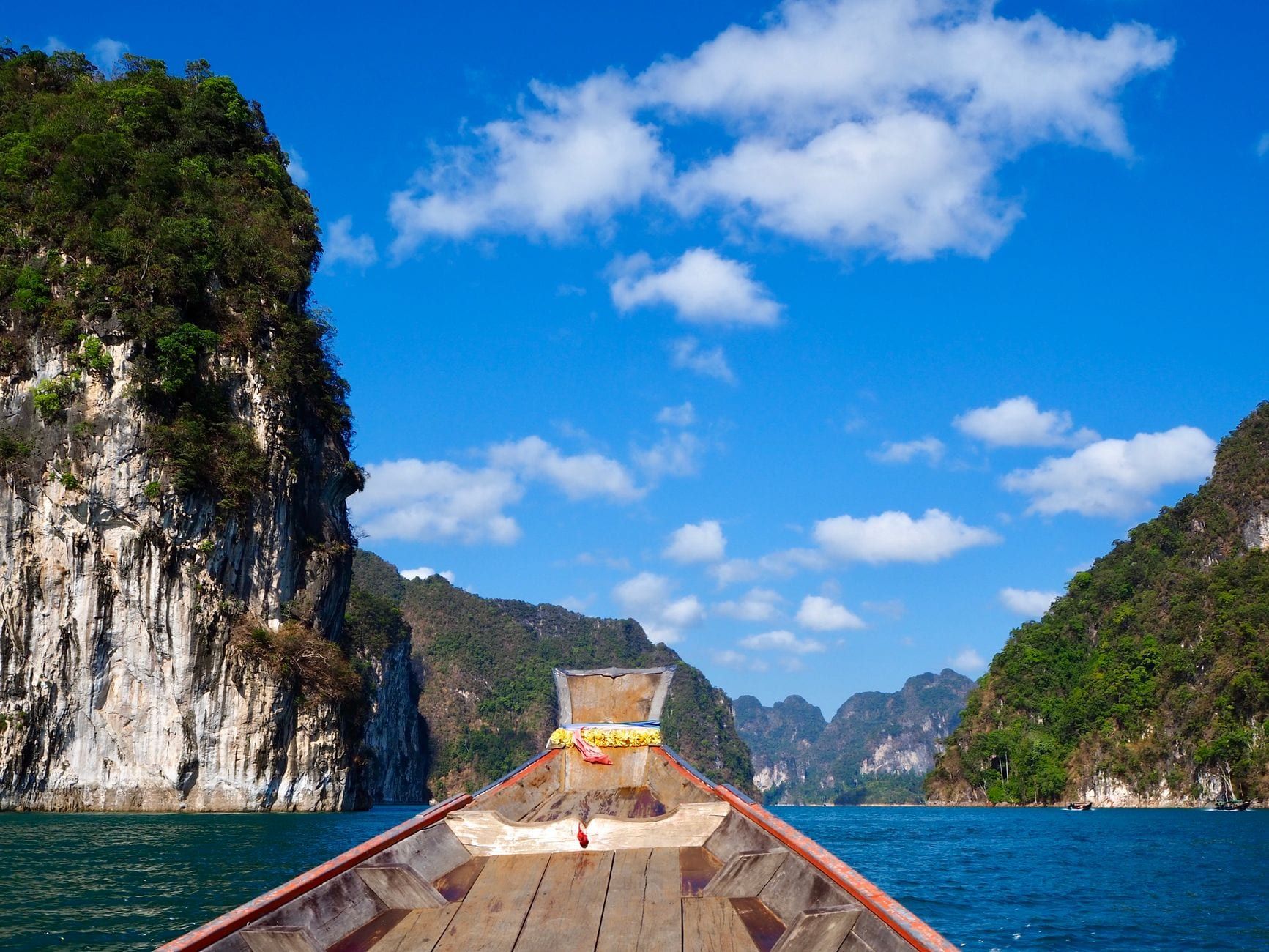 Un bote tradicional de madera navega por las vibrantes aguas azules del lago Cheow Lan en Khao Sok, bajo imponentes acantilados de piedra caliza.